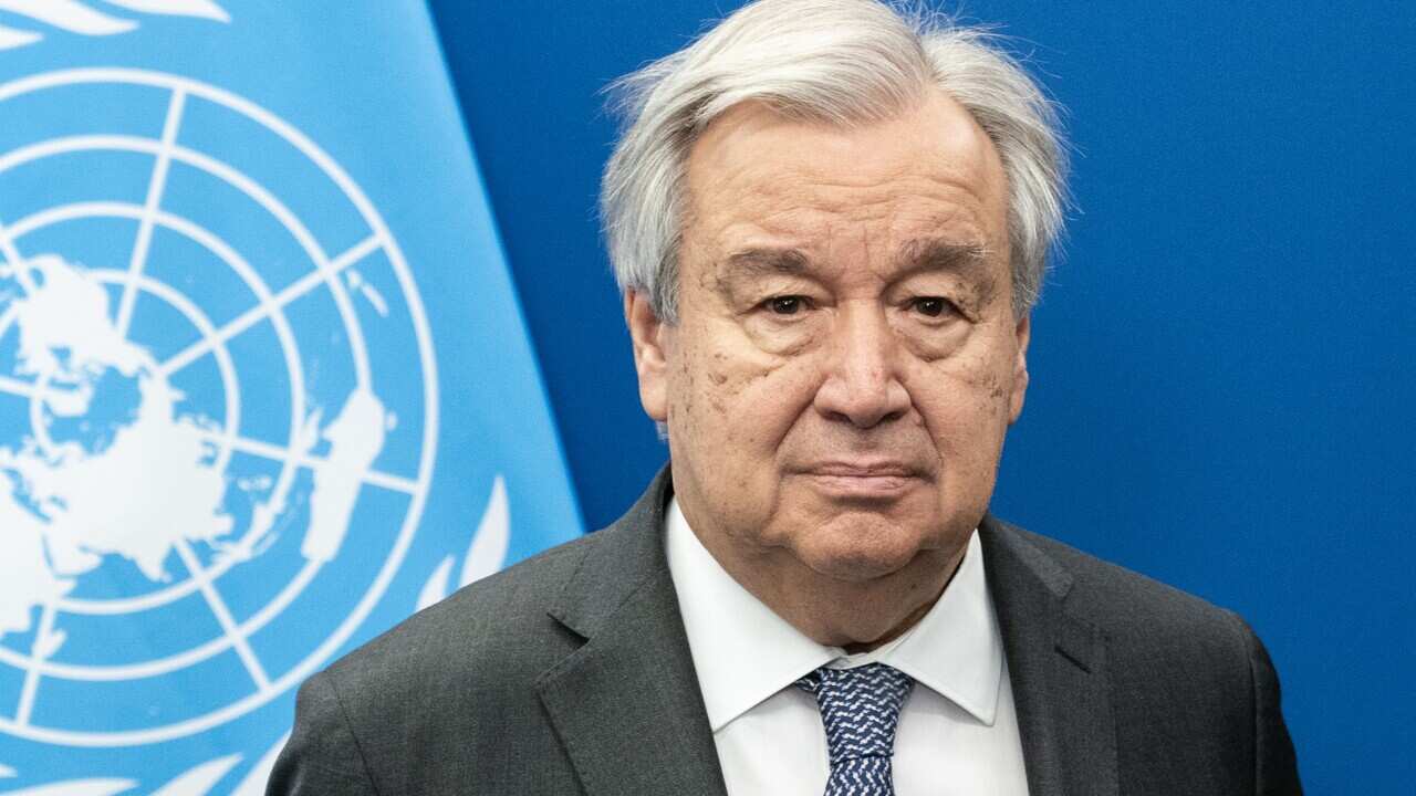 António Guterres wearing a suit standing in front of a United Nations flag