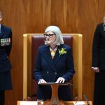 A woman with short white hair stands at a podium in the Senate chamber. Anthony Albanese is sitting to her left, while another man sits to her right and two women stand behind her