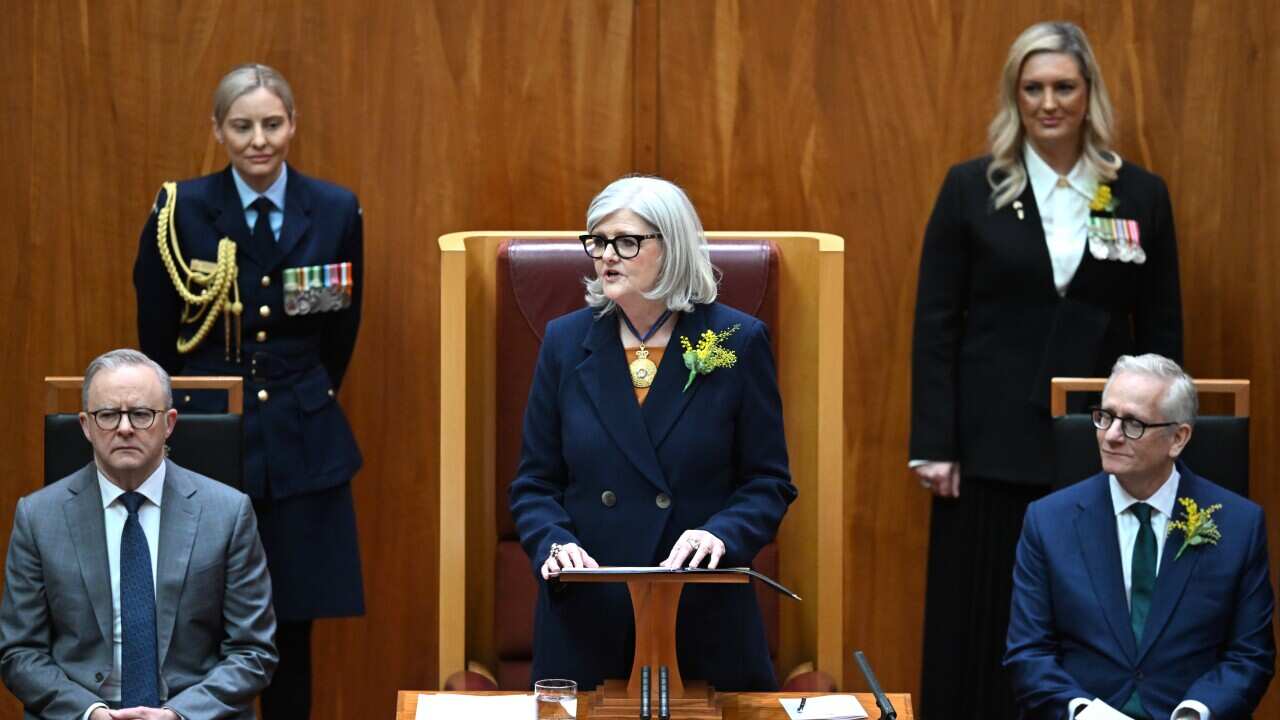 A woman with short white hair stands at a podium in the Senate chamber. Anthony Albanese is sitting to her left, while another man sits to her right and two women stand behind her