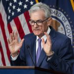 An elderly man with grey hair wearing a suit stands at a lectern speaking into two microphones. Behind him several United States flags are draped.