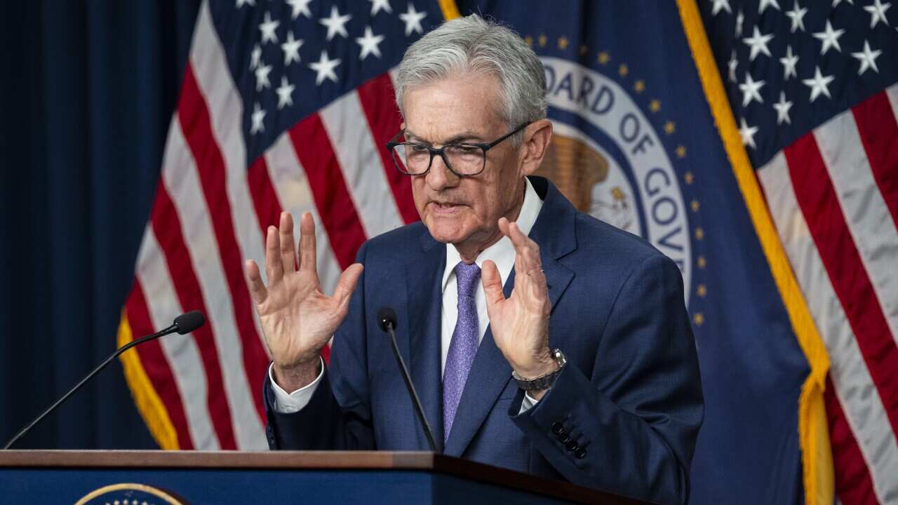 An elderly man with grey hair wearing a suit stands at a lectern speaking into two microphones. Behind him several United States flags are draped.