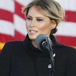 A woman in a dark coat stands at a microphone in front of the US flag