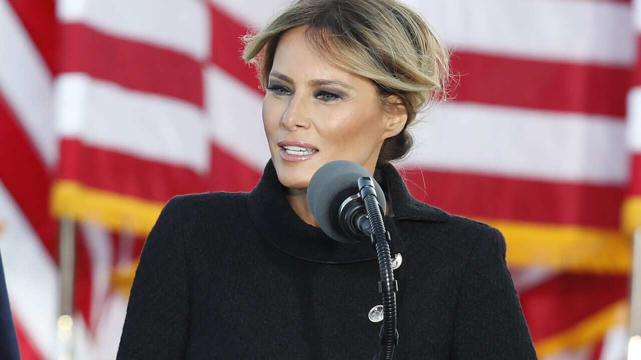 A woman in a dark coat stands at a microphone in front of the US flag