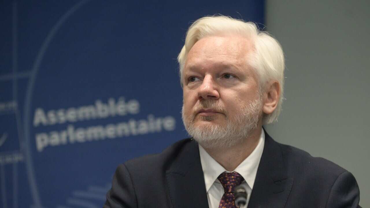 Julian Assange in a suit and tie sitting in front of a blue sign.